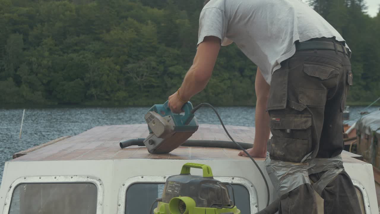 молодой устанавливает ленточную шлифовальную машину для шлифовки крыши рулевой рубки деревянной лодки