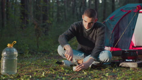 man seated on ground uses table knife to open can food near pot on fire water can in background camping outdoor activity in forest nature environment during travel leisure trip