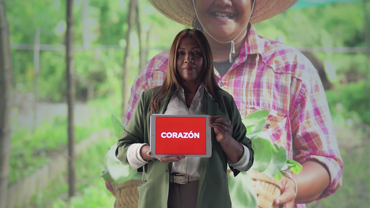 Zoom-in of a woman holding a “Corazón” sign, with background transitions showing nature, agriculture, love and care, conveying warmth and connection.