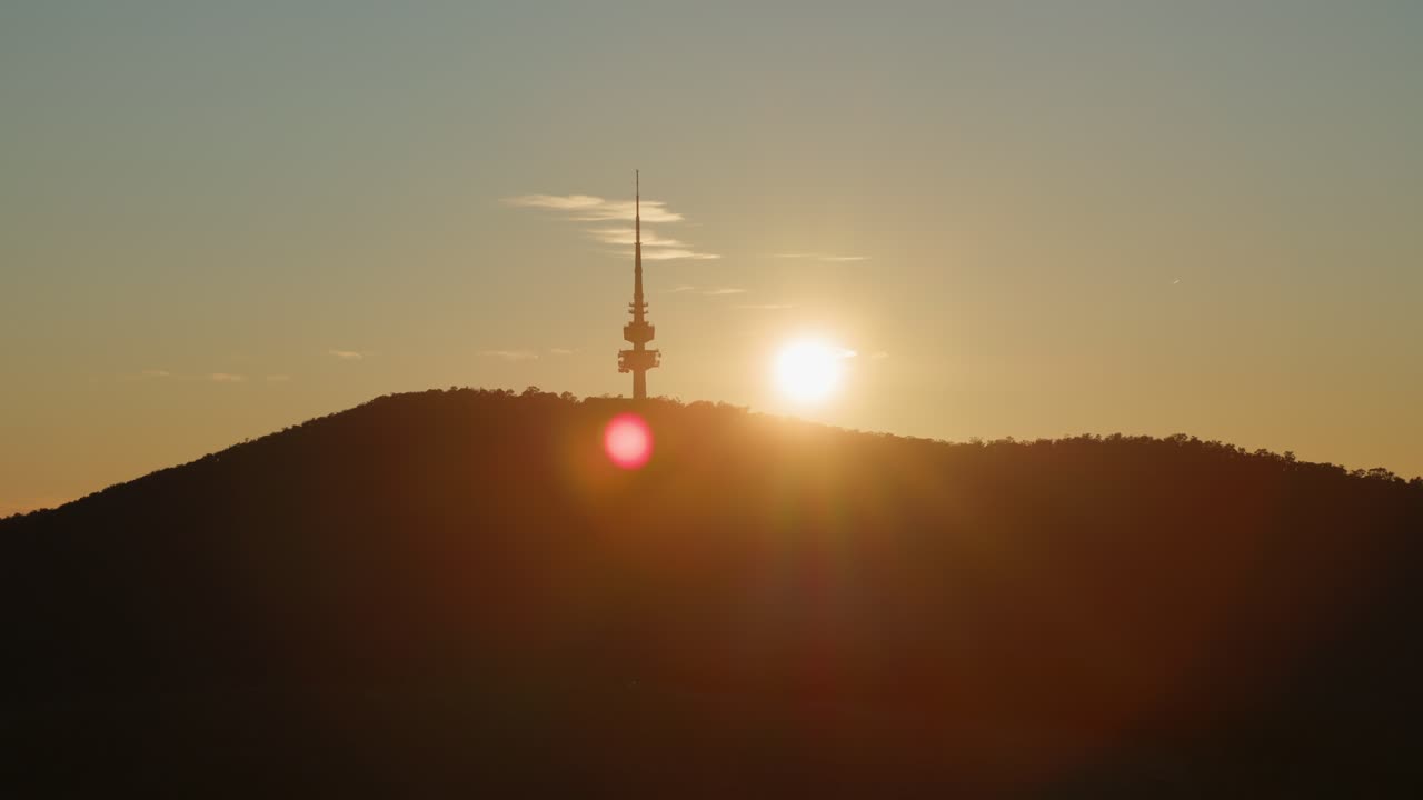 Bright morning sun rises over Black Mountain, casting a golden glow behind the silhouetted Canberra Tower in a calm start to the day