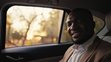 Portrait-of-a-Happy-man-with-Black-skin-and-a-beard-in-a-brown-suit-and-a-white-shirt-who-rides-in-the-passenger-seat-during-his-business-trip-in-a-modern-car-outside-the-city-during-the-sunny-day