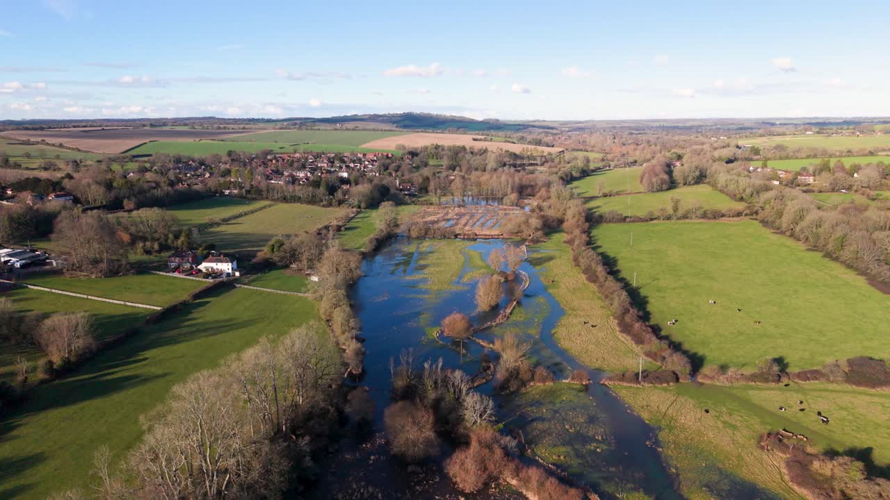 Ripresa aerea che si allontana sopra un fiume e campi allagati nella gloriosa campagna inglese nel Parco Nazionale di South Downs sotto il sole pomeridiano con ampie vedute su colline distanti e cielo blu