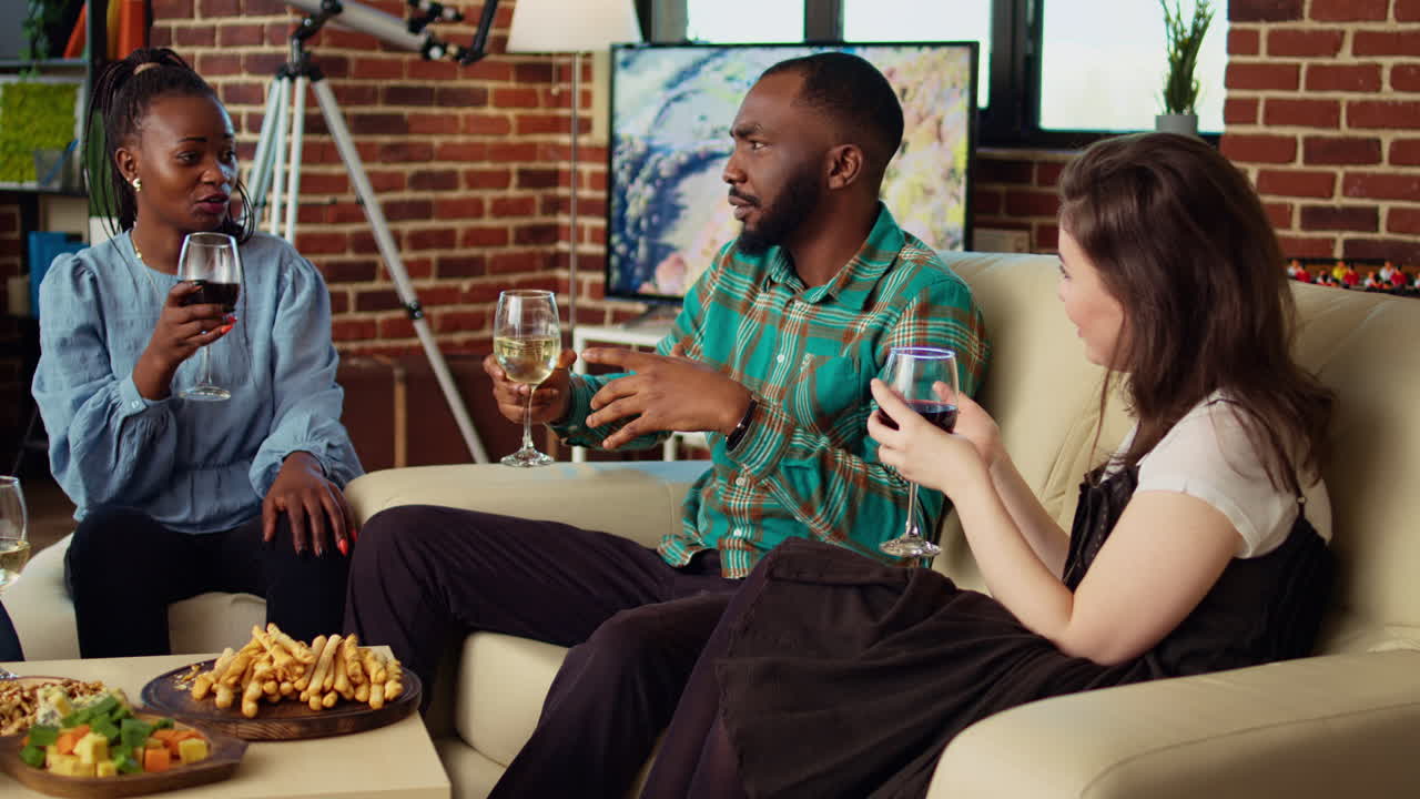 Guest making multiracial people laugh in cozy house while drinking alcohol and eating snacks