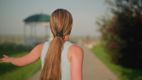 back view of young lady roller skating outdoors on sunny day, arms stretched out for balance, surrounded by lush greenery and blurred gazebo in background