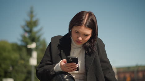 close-up of young woman in black coat and white sweater, sitting on bench outdoors, focused on smartphone screen in sunny day, bright sunlight illuminates her face and the background greenery