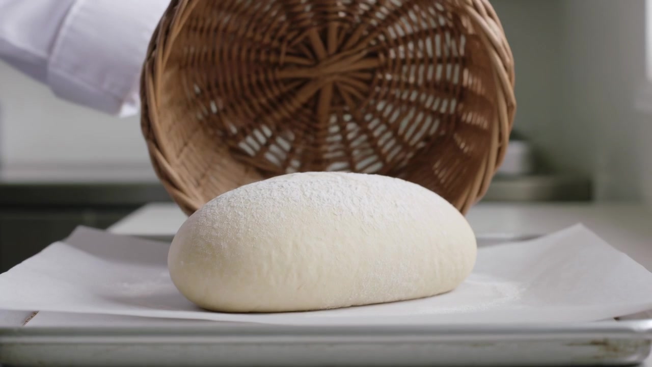 Lifting wicker basket, baker wearing white cuff revealing proofed loaf on parchment tray for baking