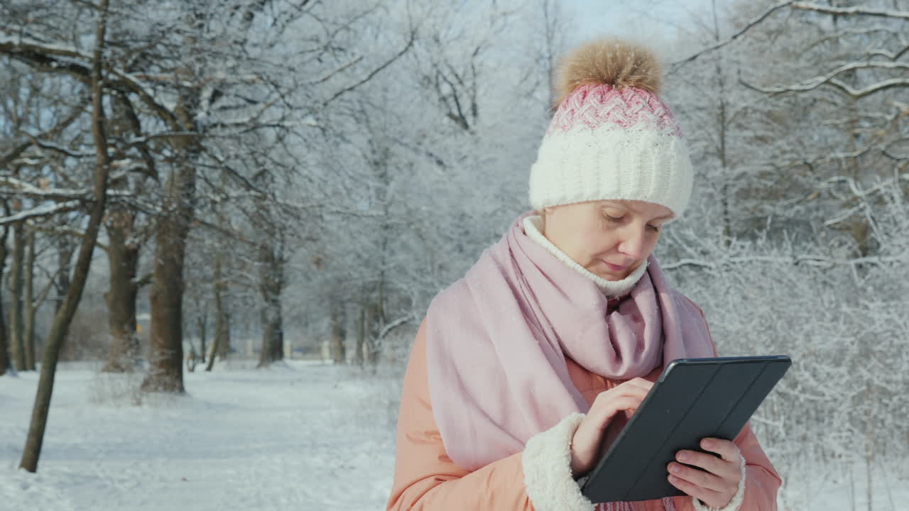 분홍색 재킷을 입은 여성이 디지털 태블릿을 사용하여 겨울 공원에서 산책을 즐깁니다.