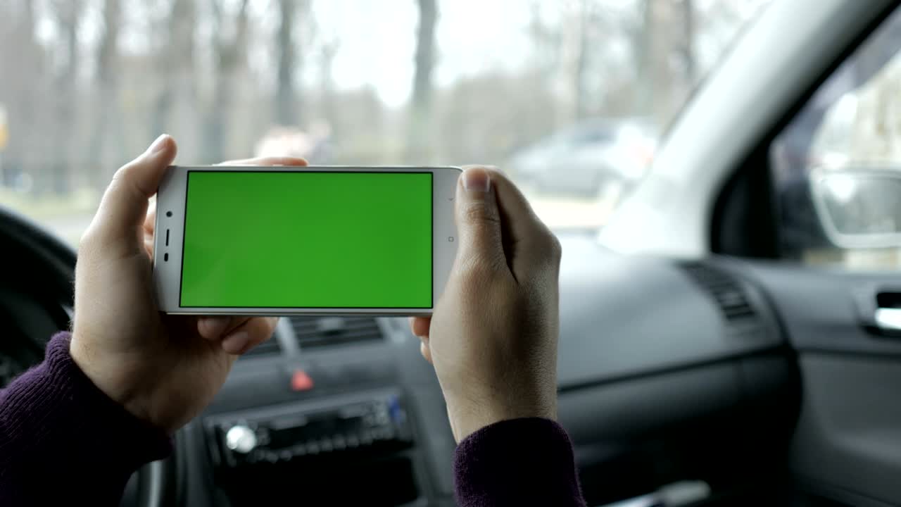 una mano sosteniendo un teléfono inteligente con pantalla verde en blanco en el coche para la dirección, masaje, ubicación, negocio. hombre se sienta en un coche moderno y trabaja en teléfono inteligente - pantalla verde - primer plano. llave de croma.