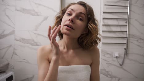 una mujer joven con una gran toalla blanca está aplicando crema en la cara bajo el área de los ojos con movimientos de masaje y mirando al espejo en el baño doméstico después de la ducha. cámara lenta