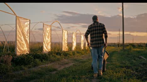 Rückansicht-Eines-Selbstbewussten-Bauern-In-Einem-Karierten-Hemd,-Der-Auf-Einem-Bauernhof-An-Einem-Feld-Entlang-Geht-Und-Abends-Bei-Sonnenuntergang-Eine-Schubkarre-Rollt