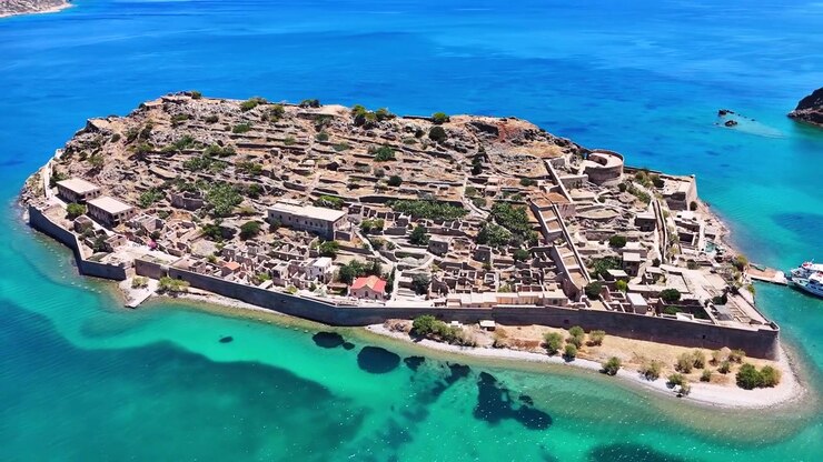 L'insediamento in rovina di Kalydon si ingrandisce in una vista aerea ravvicinata al largo di Creta, rivelando fondamenta in pietra, un bastione circolare, un muro perimetrale, alberi e un fondale scuro in acque basse turchesi
