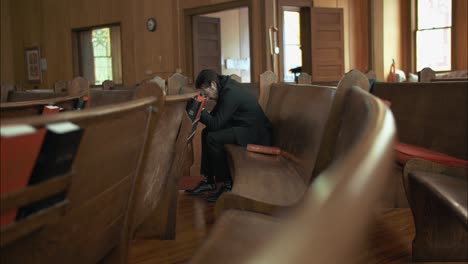 young man in black suit praying in old church pew in cinematic slow motion with folded hands