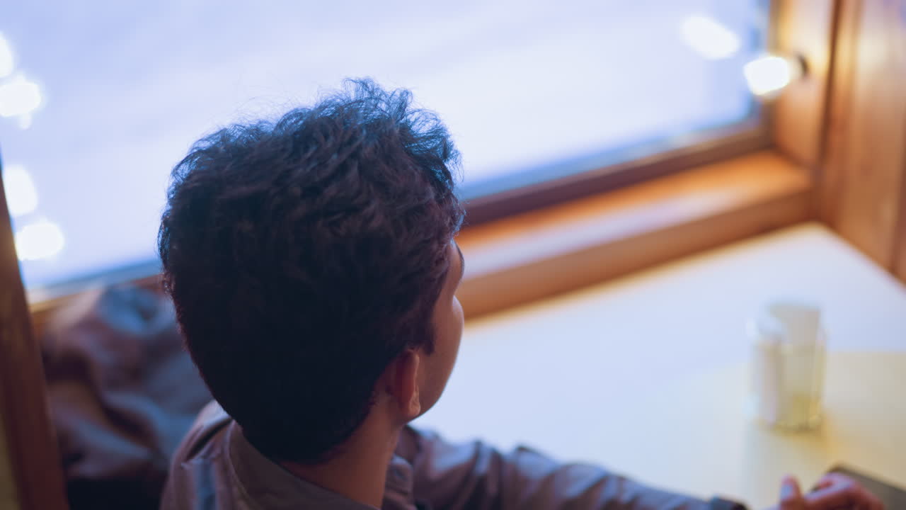 Young man sitting alone at round table near window with hands clasped, looking contemplative and distant, phone placed beside paper, evoking mood of waiting, anticipation, or quiet reflection in cozy cafe