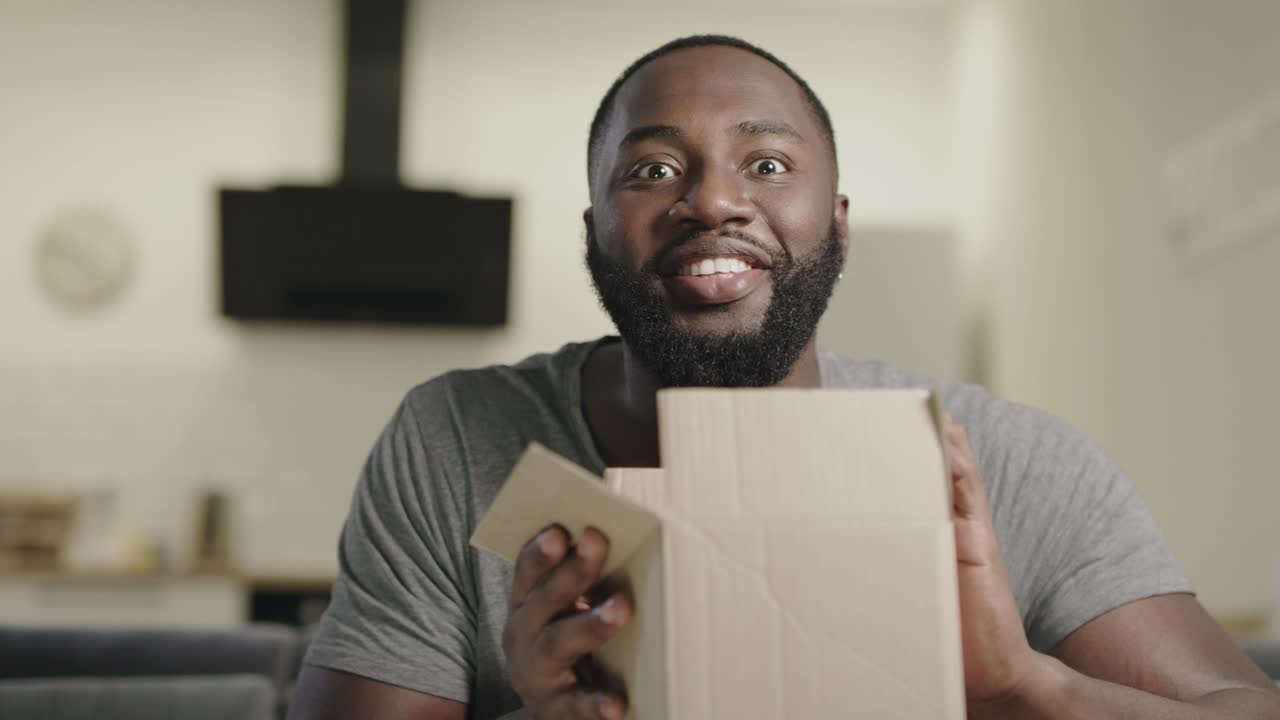 Happy black man sitting on couch with open box. Surprised young guy holding box.