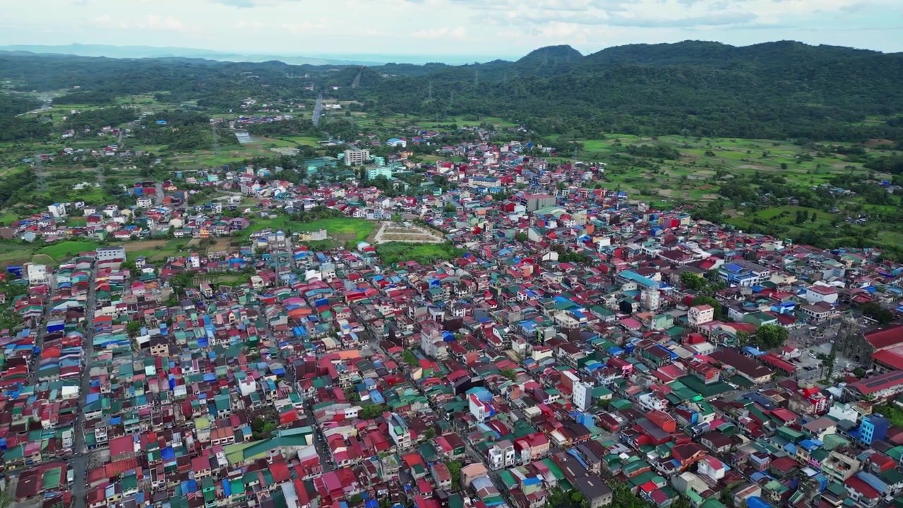 Bingkai udara yang menangkap Jalan Lingkar Lucban dikelilingi oleh rumah-rumah penduduk, sawah hijau, pohon kelapa, dan siluet Gunung Banahaw de Lucban yang mengesankan di Lucban, Quezon, Filipina