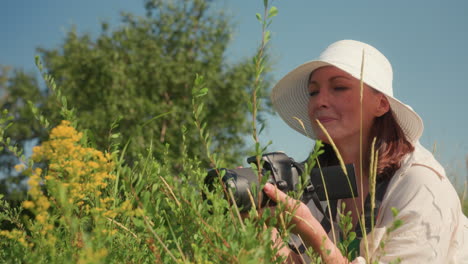 female tourist in white sunhat squats among green plants with camera in hand smiling gently while preparing to photograph wildflowers in open meadow on bright sunny day