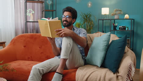 indian man relaxing interesting book turning pages smiling enjoying literature taking a rest on sofa