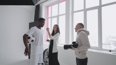 A-female-make-up-artist-makes-makeup-at-a-professional-photo-shoot-of-a-football-player.-Photo-studio-with-professional-equipment-and-team-of-workers.-team-discussing-direction-for-production