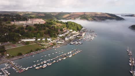 Ein-Drohnenschuss,-Der-über-Den-Fluss-Fliegt,-Zeigt-Flussaufwärts-Auf-Das-Royal-Naval-College-Von-Britannia-Mit-Einem-Nebligen-Bach-Dahinter-Und-Nebel-Weiter-Flussaufwärts