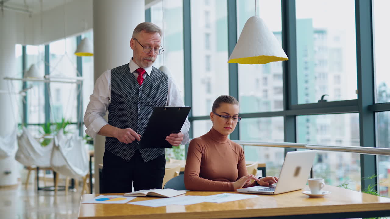 Mature man and a young woman in the office. Businessman coming to the secretary who sitting at desk and working on a laptop. Business concept.