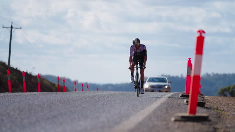 Professional-Cyclist-Riding-Bike-Uphill-Road-In-Front-Of-Car,-4K-Slow-Motion