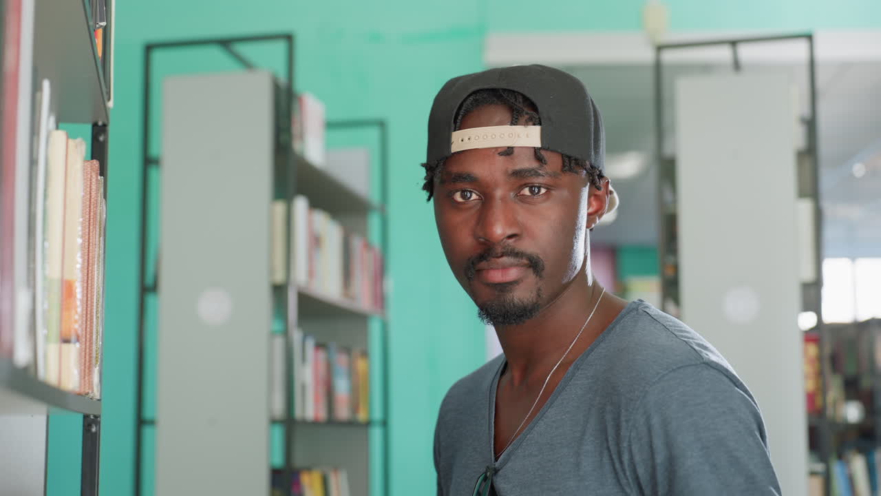 Man in cap browsing library shelf, walking between stacks, hand skimming spines, pausing to turn toward camera with focused expression, casual shirt and necklace