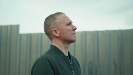 a close view of a man in a green jacket looking up thoughtfully while standing next to a corrugated metal fence