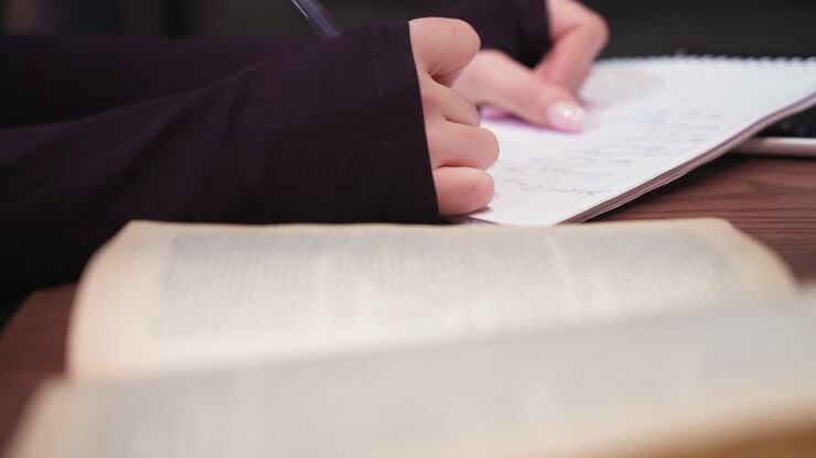 Close up view of woman hand writing with blue pen in notebook while sitting at desk with blurred open book in foreground. Scene captures quiet study moment with focus on writing action