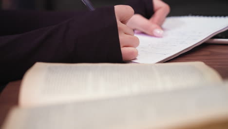 Close up view of woman hand writing with blue pen in notebook while sitting at desk with blurred open book in foreground. Scene captures quiet study moment with focus on writing action