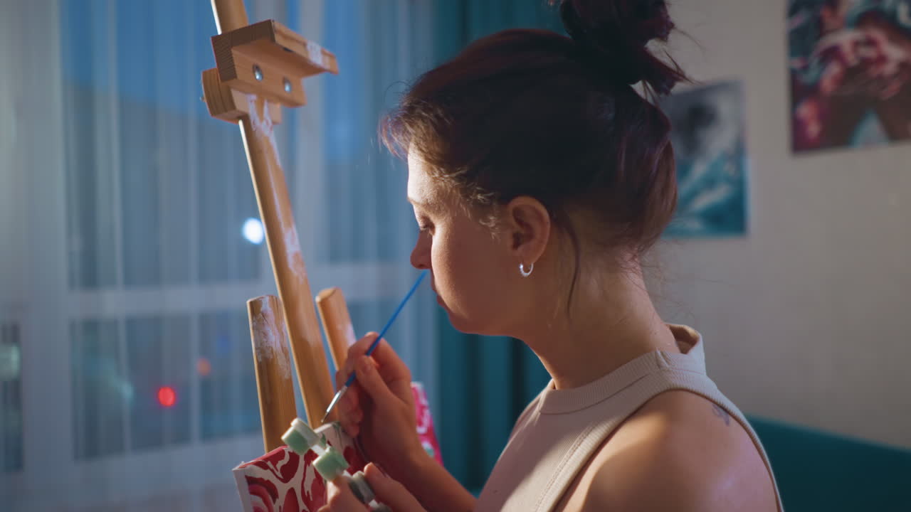 Young Caucasian Woman Painting At Easel, Cozy Apartment Studio, Closeup On Hands And Brush, Soft Window Light And Candle Glow, Thoughtful Expression While Mixing Pink Tones On Palette, Evening