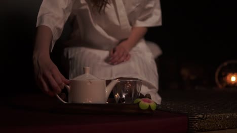 Beautiful-brunette-in-bathrobe-taking-teapot-and-pouring-tea-in-cup-drinking-herbal-tea-at-the-spa-in-slow-motion.-Thai-tea