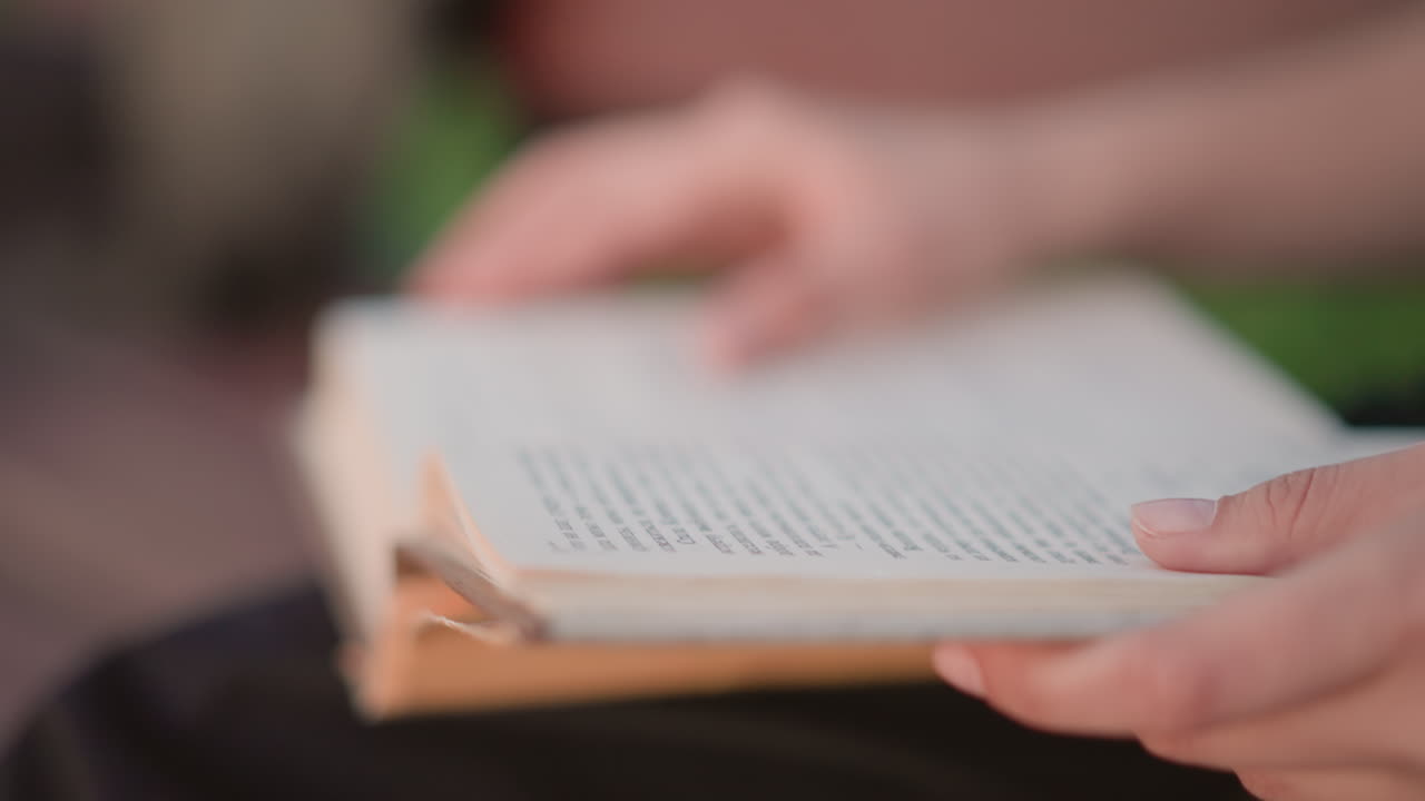 mani che sfogliano le pagine di un libro sulle gambe, luce dorata soffusa, primo piano di dita che tracciano righe su un libro tascabile usato, ambiente esterno con panchina, ritmo lento e intimo, studente che studia poesia, profondità di campo ridotta, caldo