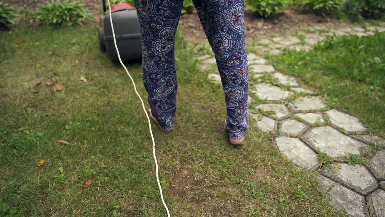 Woman's legs and corded lawn mower. Worker cutting grass in the garden. Gardening activity. Female worker pushing mowing machine.