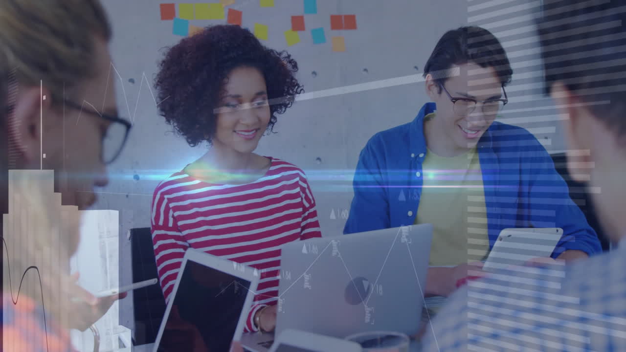 Colleagues collaborating at table, typing on laptops and tablets displaying business chart overlays