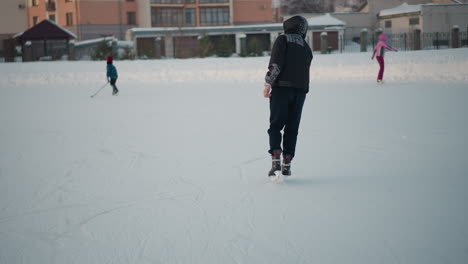 young kid skating on outdoor ice rink during winter season wearing warm clothes, enjoying winter sport in park with snow, trees, and buildings in soft background light