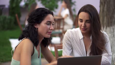 dos mujeres sentadas en la cafetería al aire libre - hablando, compartiendo noticias, chica morena mostrando algo en la pantalla de la computadora portátil, gestando