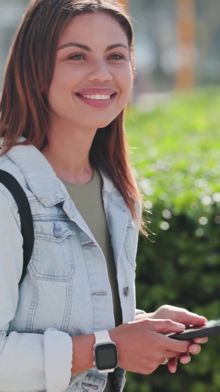 Video verticale: Donna in denim che controlla l'orologio, tocca il telefono e legge con uno zaino su un sentiero vicino a una siepe.