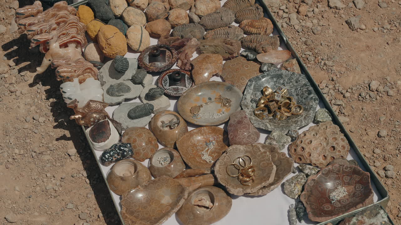 Fósiles de trilobites, piedras pulidas, anillos y figurillas talladas en una bandeja de exhibición sobre suelo desértico cerca de Erfoud.