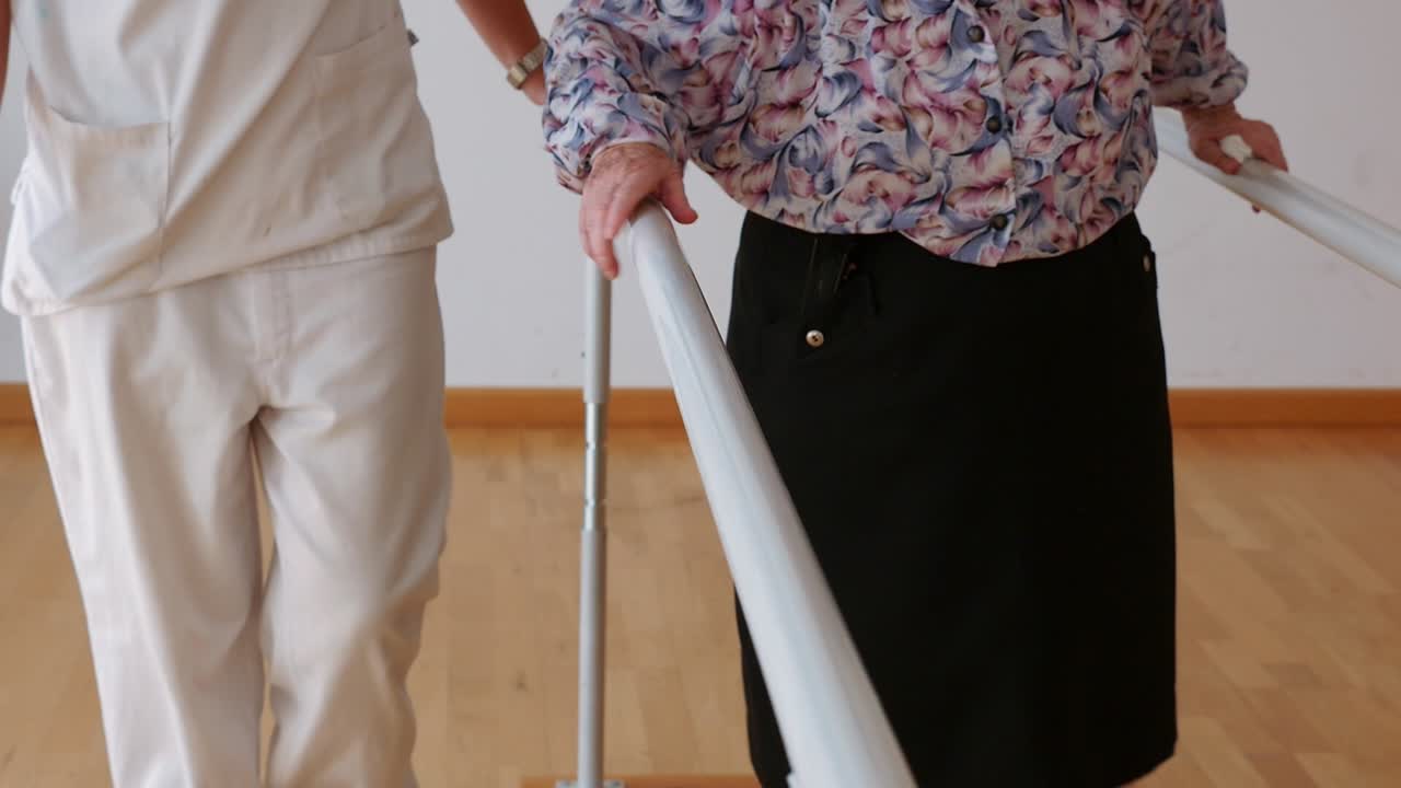 Elderly woman practices walking on parallel bars under the supervision of a physiotherapist. Leg rehabilitation, balance, and walking safety at an active geriatric center