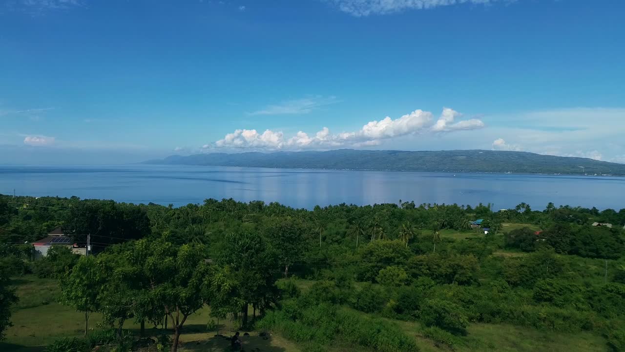 FPV flyover view of houses among lush green trees with the view of Cebu island in the expansive horizon and the blue summer sea