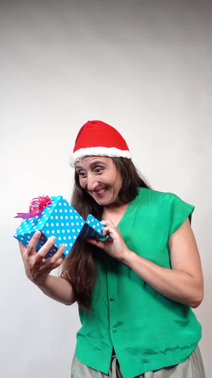 Long-haired woman in green top and red Santa hat beams, shows OK sign with wrapped blue gift