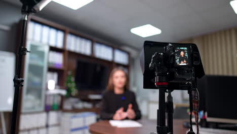 modern camera on tripod shooting a blog content. female speaker influencer sits at desk talking. blurred backdrop.