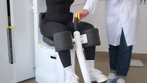 a woman is sitting on a chair that looks like sports equipment with a doctor standing behind her. the machine focuses on her leg, thigh, and knee joint