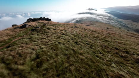 Aerial-view-of-a-mountain-ridge-covered-in-grass-and-rocks-with-clouds-below-and-clear-sky-above