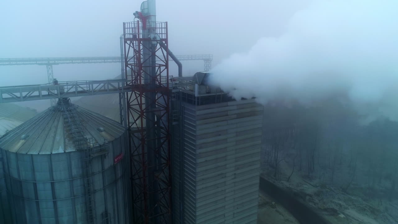 Grain processing plant premises on gray foggy day. Functioning enterprise producing heavy smoke into air. Aerial view.
