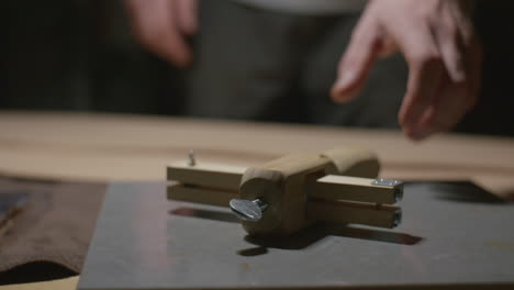 a detailed view of a wooden leather edge tool resting on a work surface, with a craftsman’s hands reaching towards it in a dimly lit workshop.