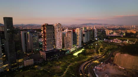 Drone-flying-in-front-of-sunlit-buildings-and-the-Parque-La-Mexicana,-sunny-evening-in-Santa-Fe,-Mexico