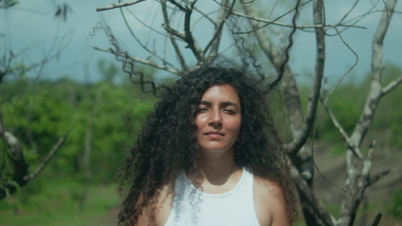 A woman stands motionless in front of a tree, her long curly hair caught in the branches as the camera pulls back