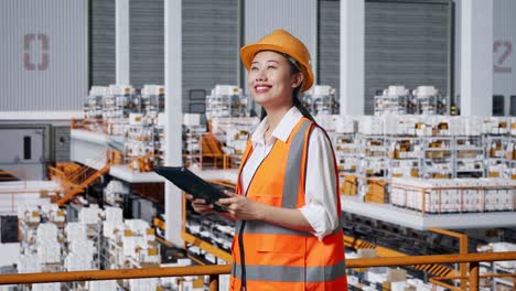 vista lateral de una ingeniera asiática con casco de seguridad mirando la tableta en su mano y mirando a su alrededor mientras está de pie en el almacén con estantes llenos de productos de entrega