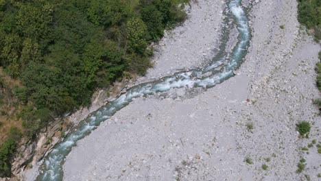 Aerial-overhead-view-captures-a-serene-river-meandering-through-a-rocky,-dry-valley-surrounded-by-lush-green-foliage,-emphasizing-the-contrast-between-nature's-differing-landscapes-and-elements